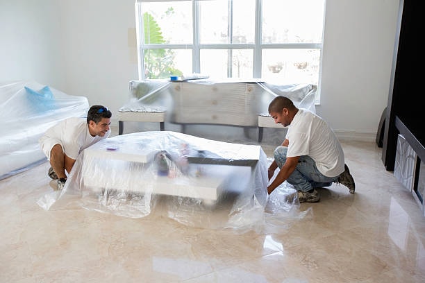 2 painters lifting up table furniture in house before starting painting. entire house furnitures are covered in plastic. 