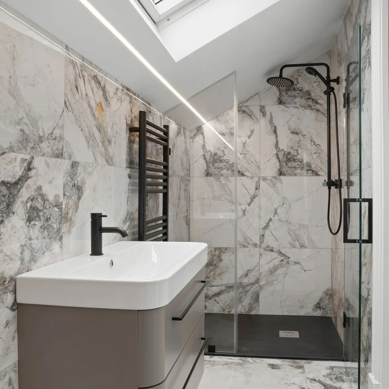 Modern en-suite with marble tiles, black rain shower, floating vanity, LED strip lighting, and skylight.