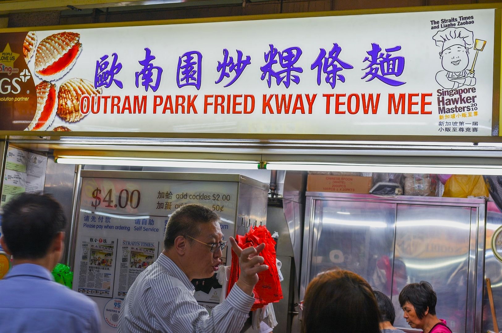 Street food stall with a bright sign reading "Outram Park Fried Kway Teow Mee" in purple and red text. People are lined up, one holding a red plastic bag. Buzzing atmosphere.