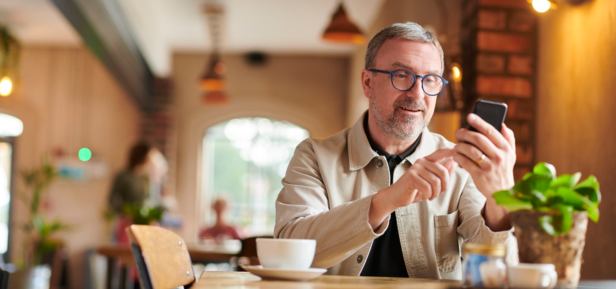 Mature man in round glasses and a beige jacket checking his email in a coffee shop.
