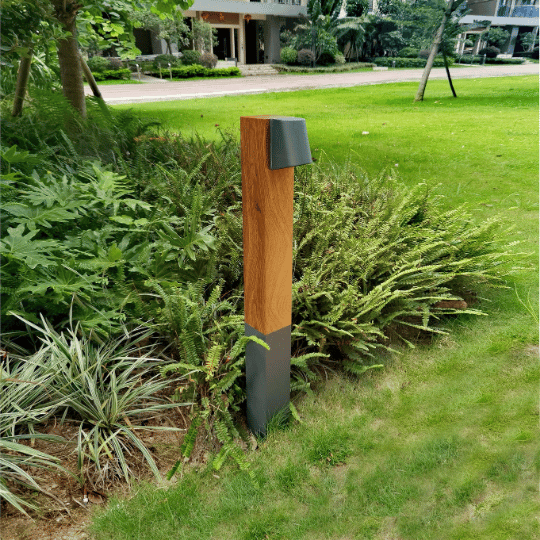Japandi-style outdoor bollard light with a wooden top and metal base, set among lush greenery in a well-maintained garden, adding soft illumination to the outdoor space.