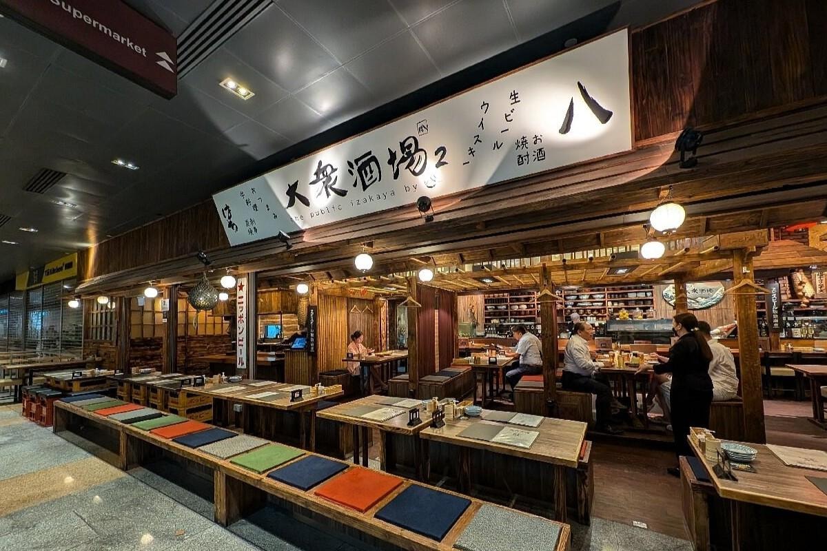  This image captures the exterior of The Public Izakaya 2, a Japanese restaurant featuring a rustic wooden design with long bench seating and colorful cushions. A large, brightly lit sign with Japanese calligraphy hangs above the entrance, while the interior reveals customers dining under warm, spherical lanterns.