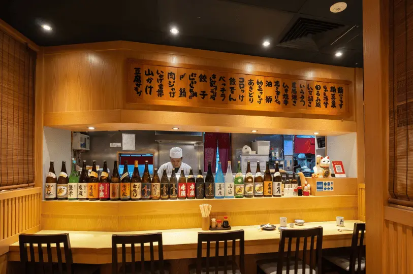 A chef working behind a wooden counter at a Japanese restaurant, with a traditional wooden menu board hanging above and a diverse row of sake bottles lining the counter in the foreground.