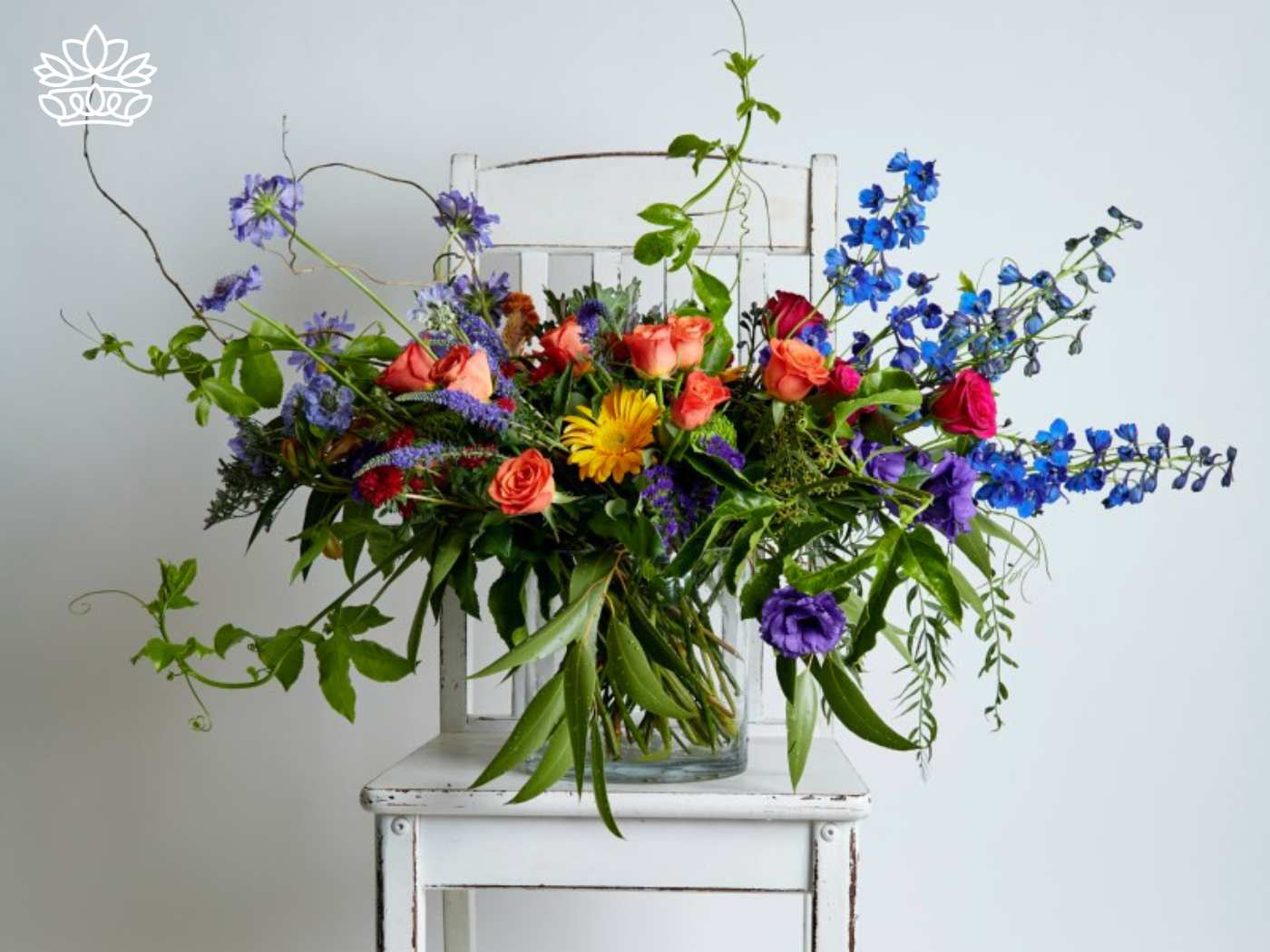 A vibrant and artistically arranged bouquet of flowers displayed on a rustic white chair, featuring an eclectic mix of roses, delphiniums, and wildflowers in a spectrum of blues, purples, and oranges, capturing the essence of bespoke elegance and freshness. Fabulous Flowers and Gifts for Guest House and Hotel Flowers"