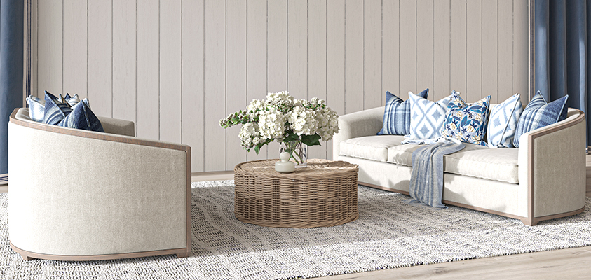 This relaxed living room is the perfect mix of natural elements that is signature to Hamptons styling. Two cream couches with blue throw pillows sit opposite a round wicker coffee table that displays flowers in a vase. A large rug breaks up the light timber flooring, and blue curtains soften the beige shiplap walls to tie the room together.