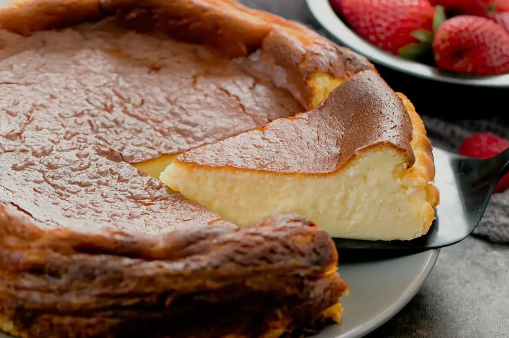 A slice of creamy cheesecake being served from a round cheesecake with a golden-brown crust, alongside fresh strawberries in the background.