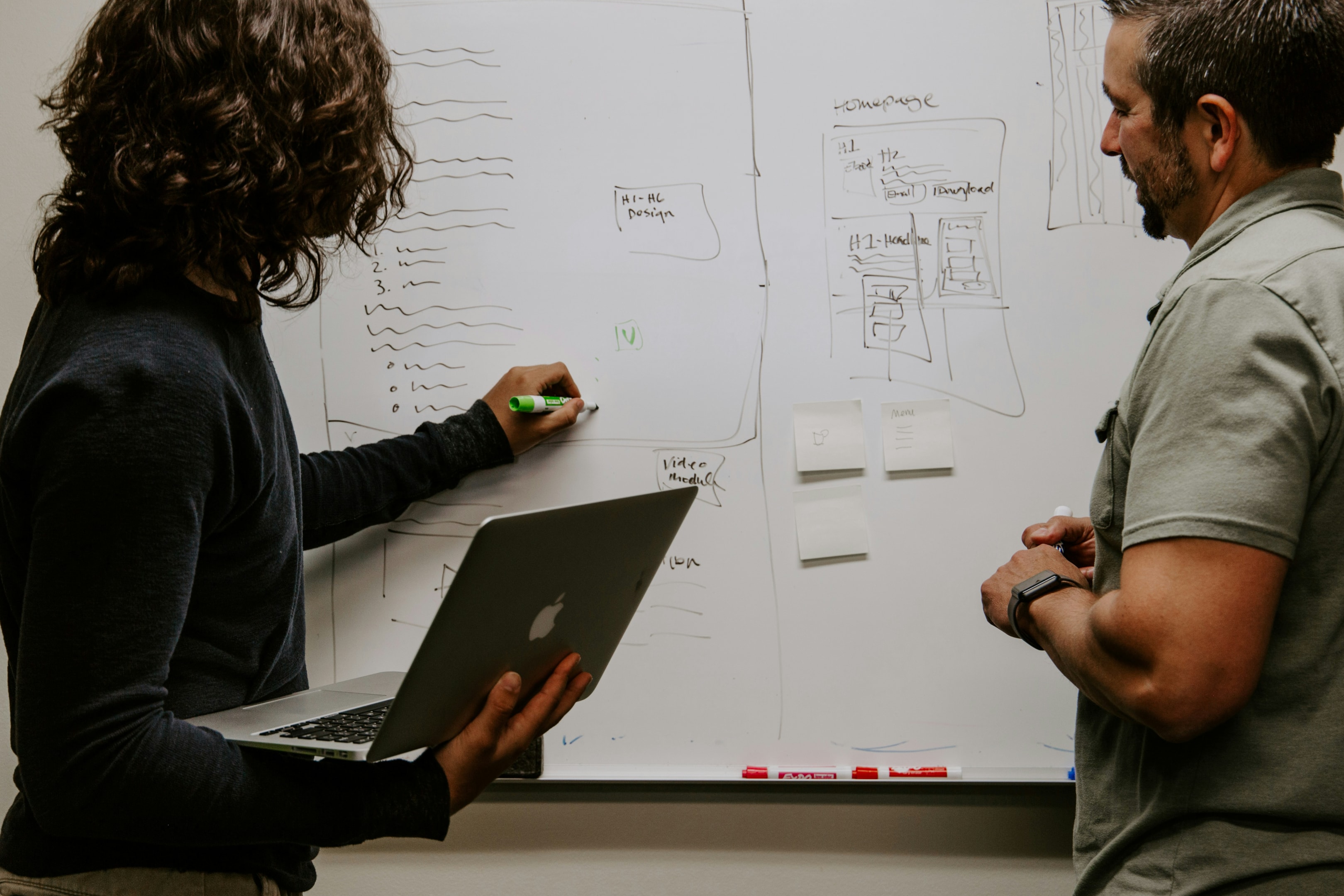 Woman writing on whiteboard, holding laptop, man standing beside her