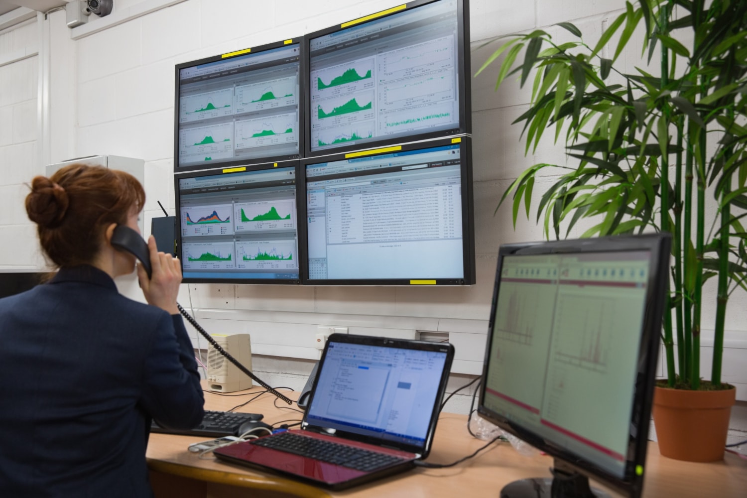A female security analyst is speaking on the phone while working at a multi-screen security station filled with data and monitoring feeds. The cloud-based surveillance system aids in workplace security and monitoring secure office entry.