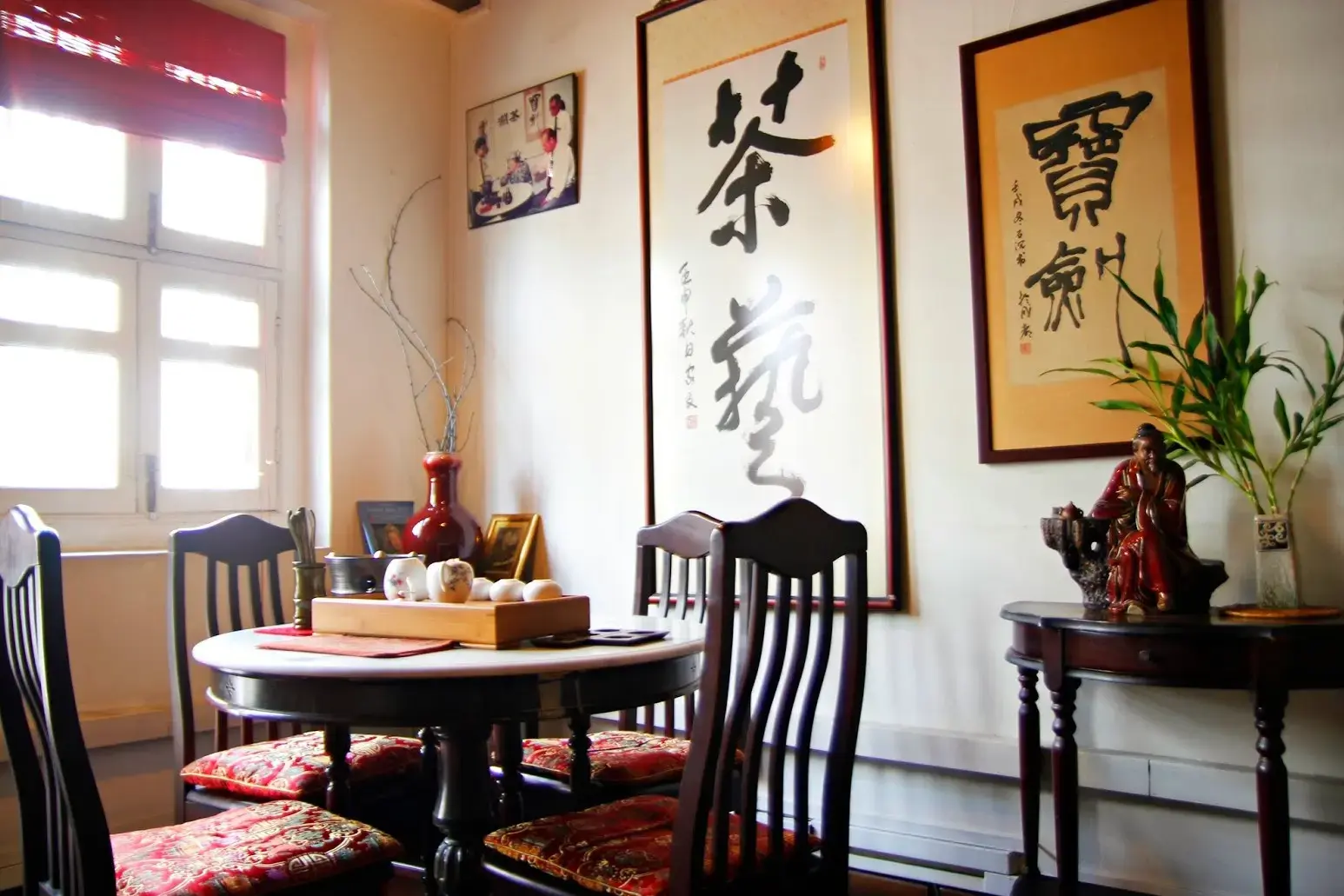 This serene indoor setting features a traditional Chinese tea room with dark wooden furniture and vibrant red cushions. Elegant calligraphy scrolls and a decorative tea set on the table create a tranquil, culturally rich atmosphere.