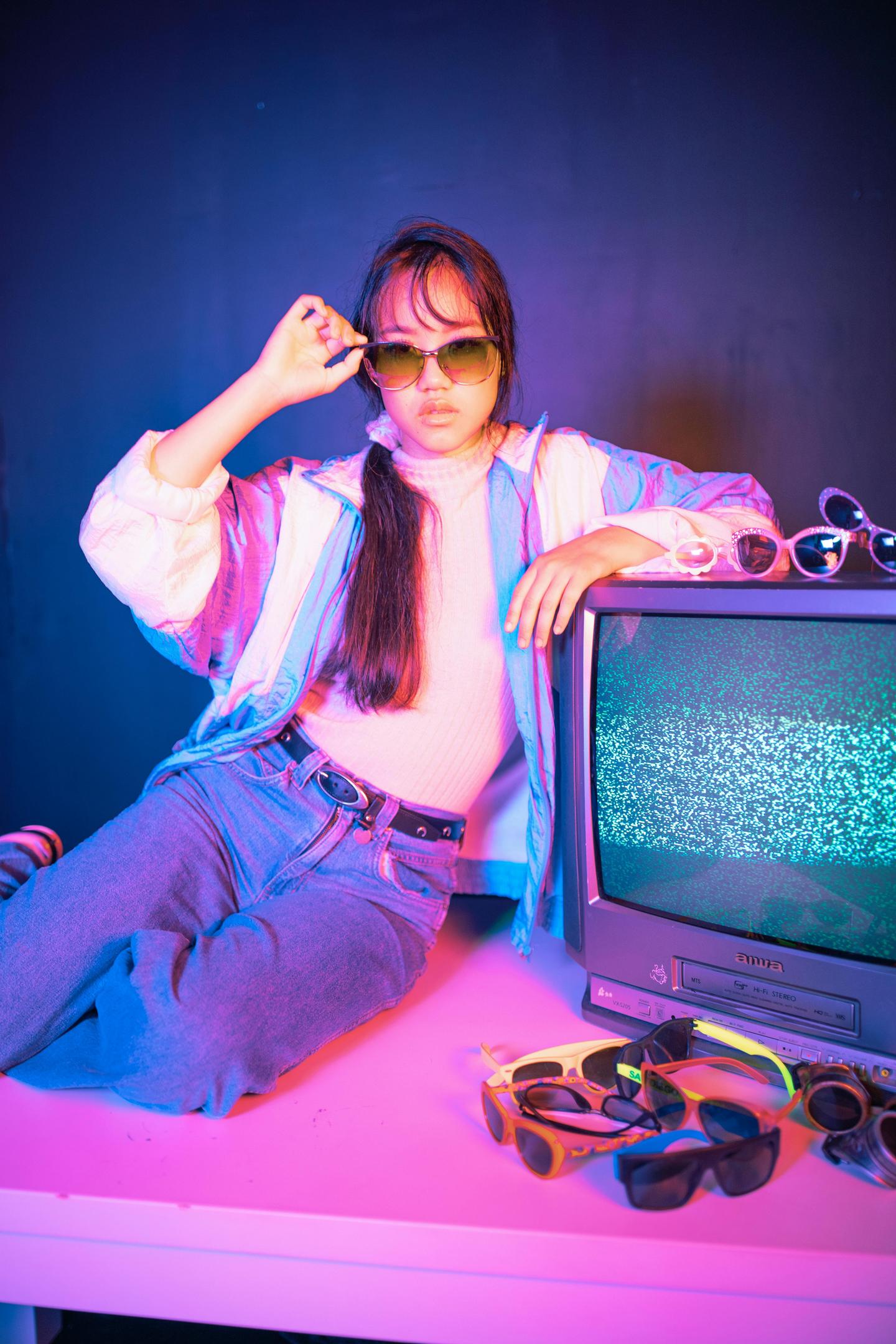 Young woman dressed in 80's style clothing including blue, straight leg jeans, a windbreaker and tortoise shell sunglasses.