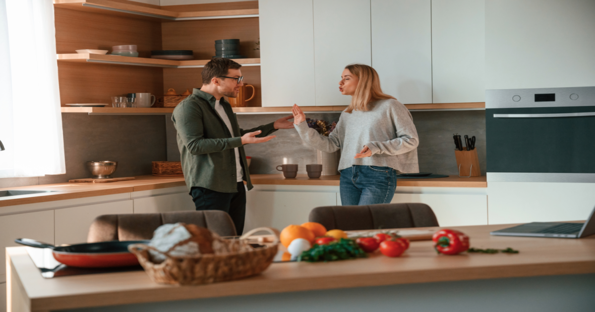 A photograph of a couple standing in a kitchen, with one partner looking upset and the other appearing defensive. The couple appears to be in the midst of an argument or disagreement, possibly related to household chores or responsibilities. The image conveys the idea of a couple who may be experiencing difficulties in their relationship and could benefit from the services of a certified Gottman Method Couples Therapist in New York City. The therapist can help the couple to develop effective communication skills, build emotional connection, and reduce conflict in their relationship, even in everyday situations like household chores.