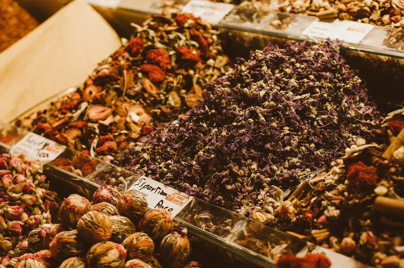 A vibrant, eye-level close-up displays a variety of dried herbs and flowers for tea organized in clear, rectangular bins at a market. The foreground is dominated by a large mound of deep purple, dried flower buds, while other compartments contain assorted dried fruits, petals, and tightly rolled flower tea balls, labeled with handwritten tags.