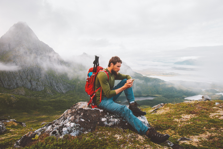 Young man carrying a red backpack sitting on a tree stump sending a text.