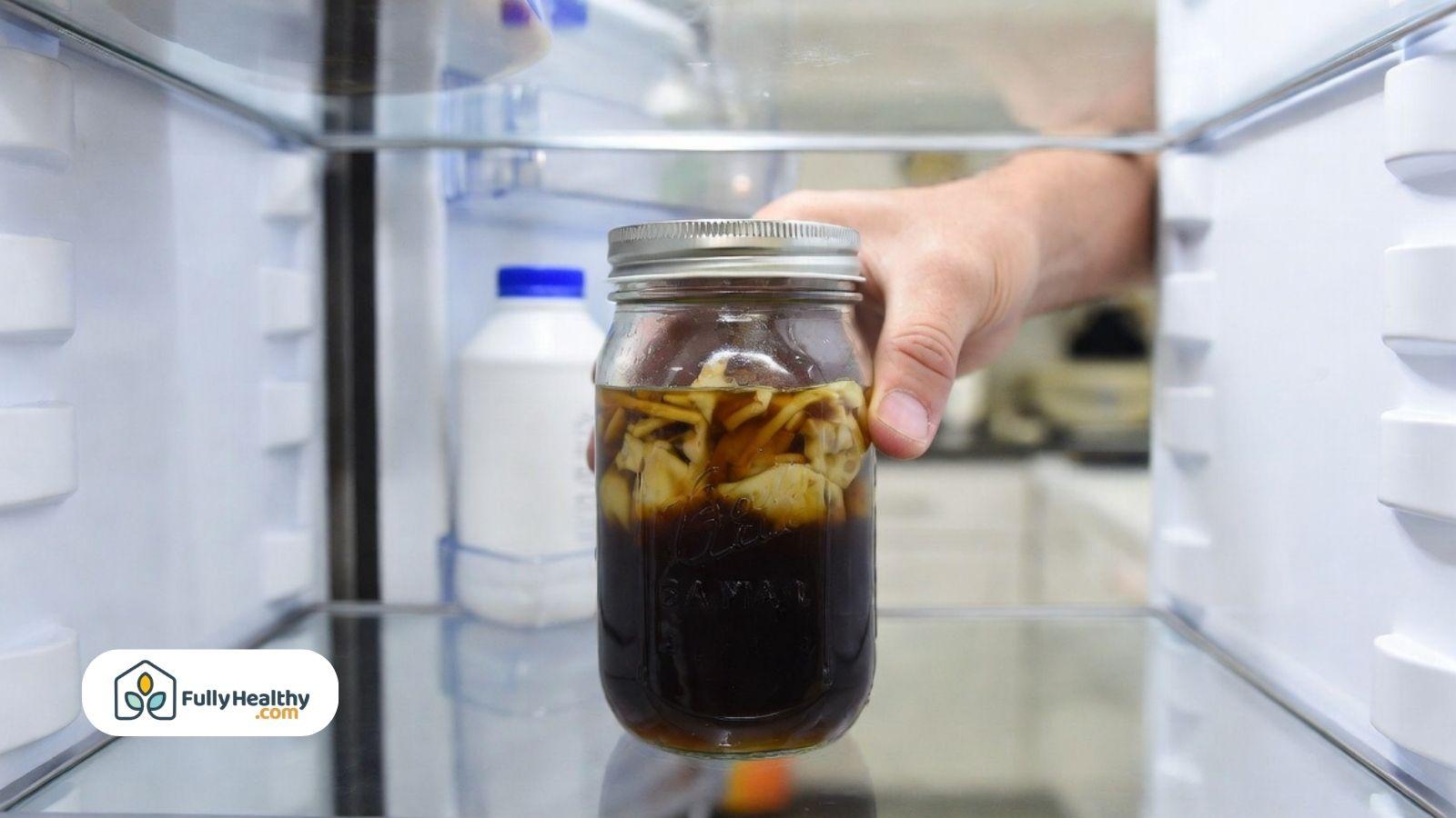 Jar of dark broth being placed inside a refrigerator shelf by hand.
