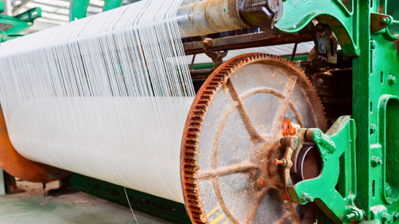 Close-up of a loom with white threads on a large spool and a rusty gear mechanism.