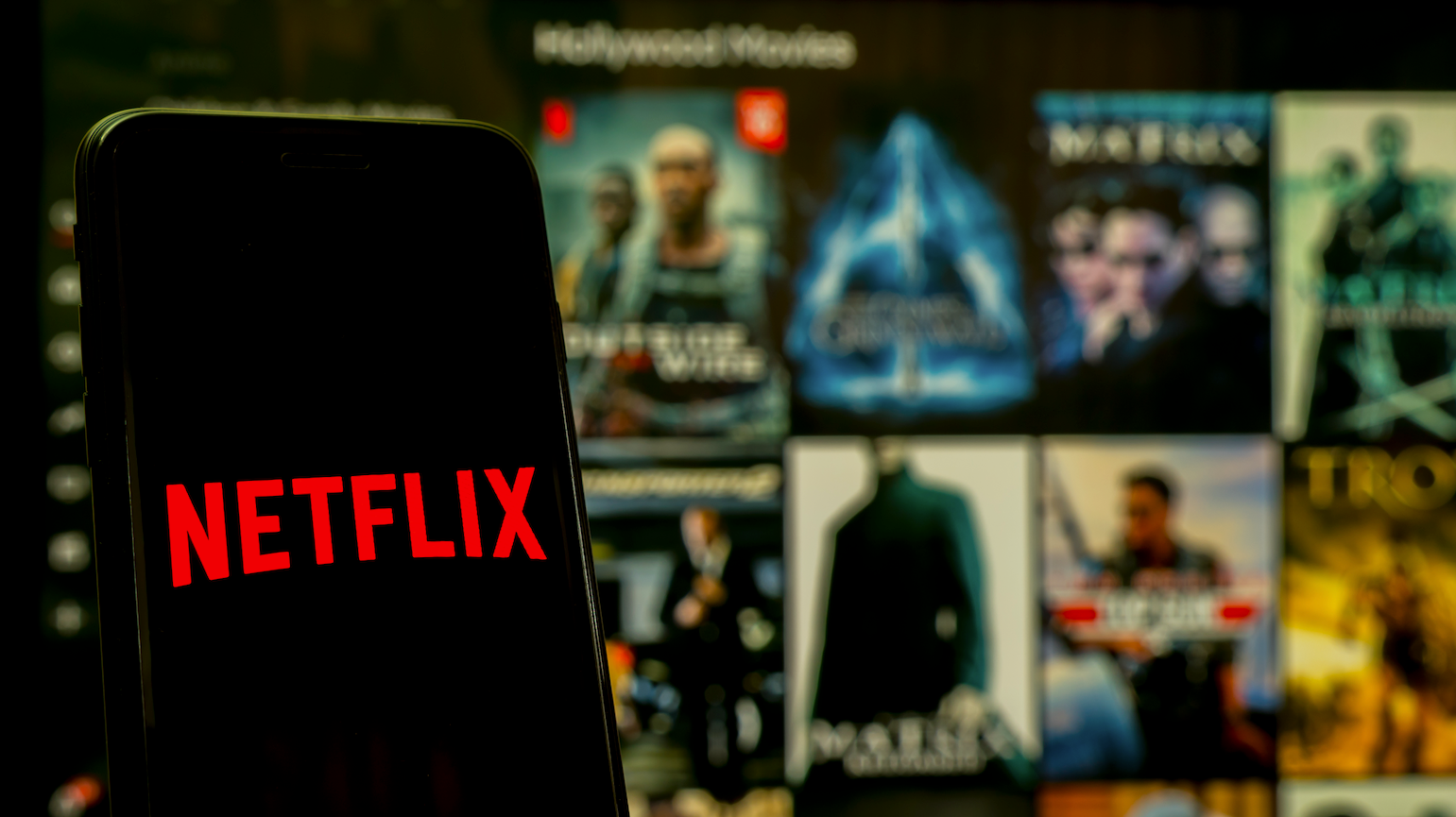 a phone displays the netflix logo against a TV screen showing movies to choose from