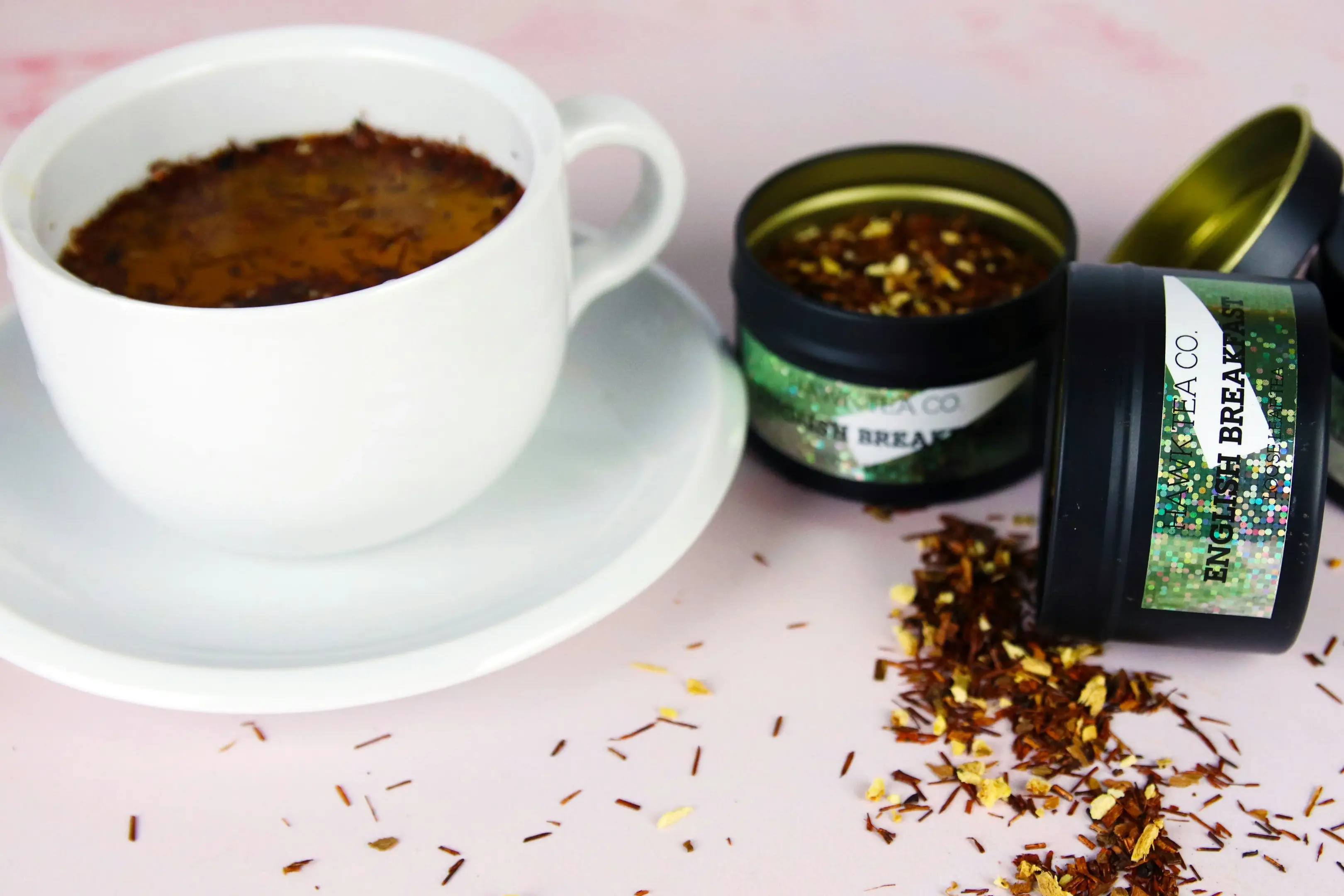 A white ceramic teacup filled with dark amber tea sits on a matching saucer alongside several black tins of loose-leaf English Breakfast tea. Loose tea leaves are scattered across the light pink surface, creating a textured and aromatic scene.