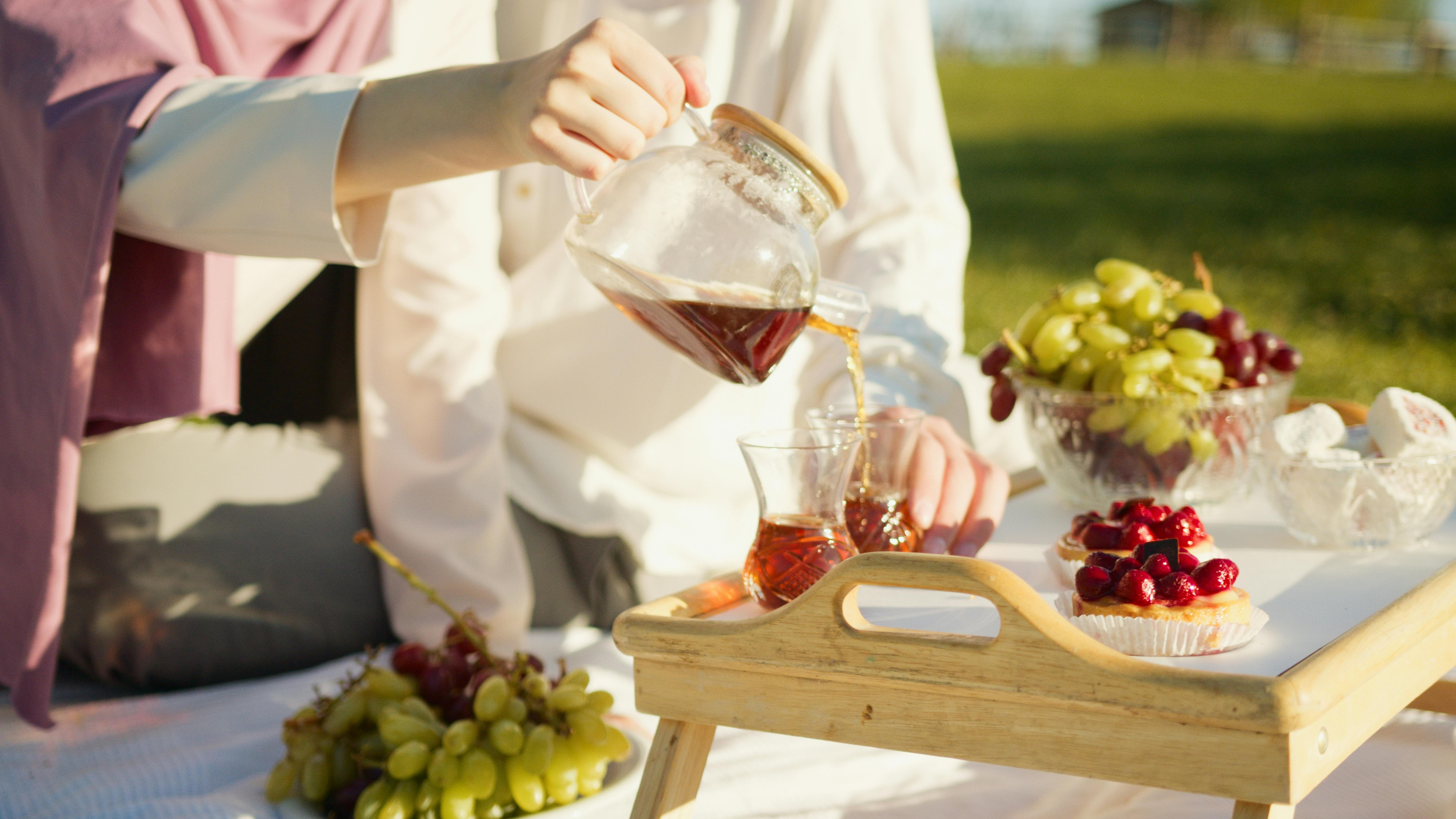 person pouring coffee into clear glass, grapes and pastries on wooden tray