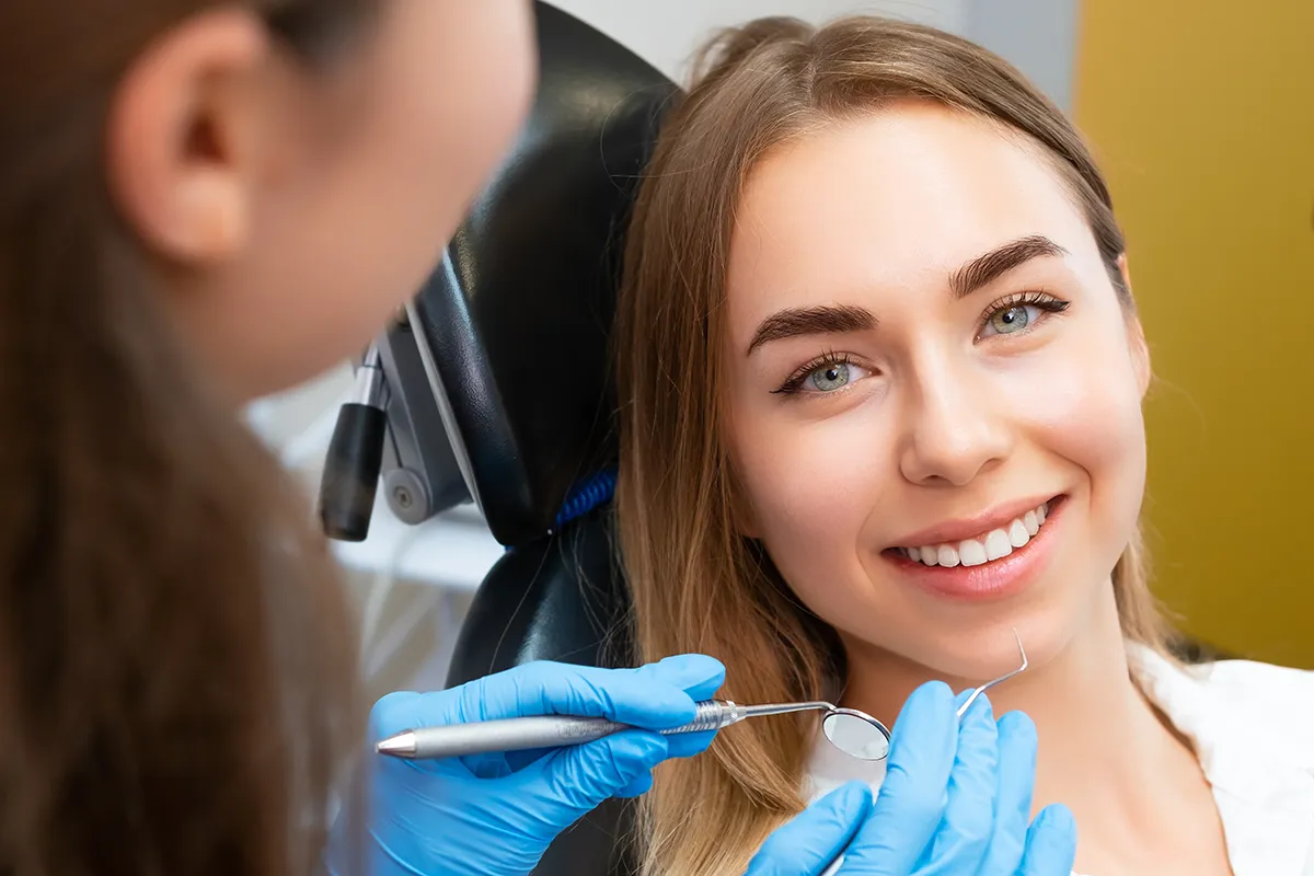 A smiling female patient in a dental chair as a dentist with blue gloves prepares to examine her teeth.