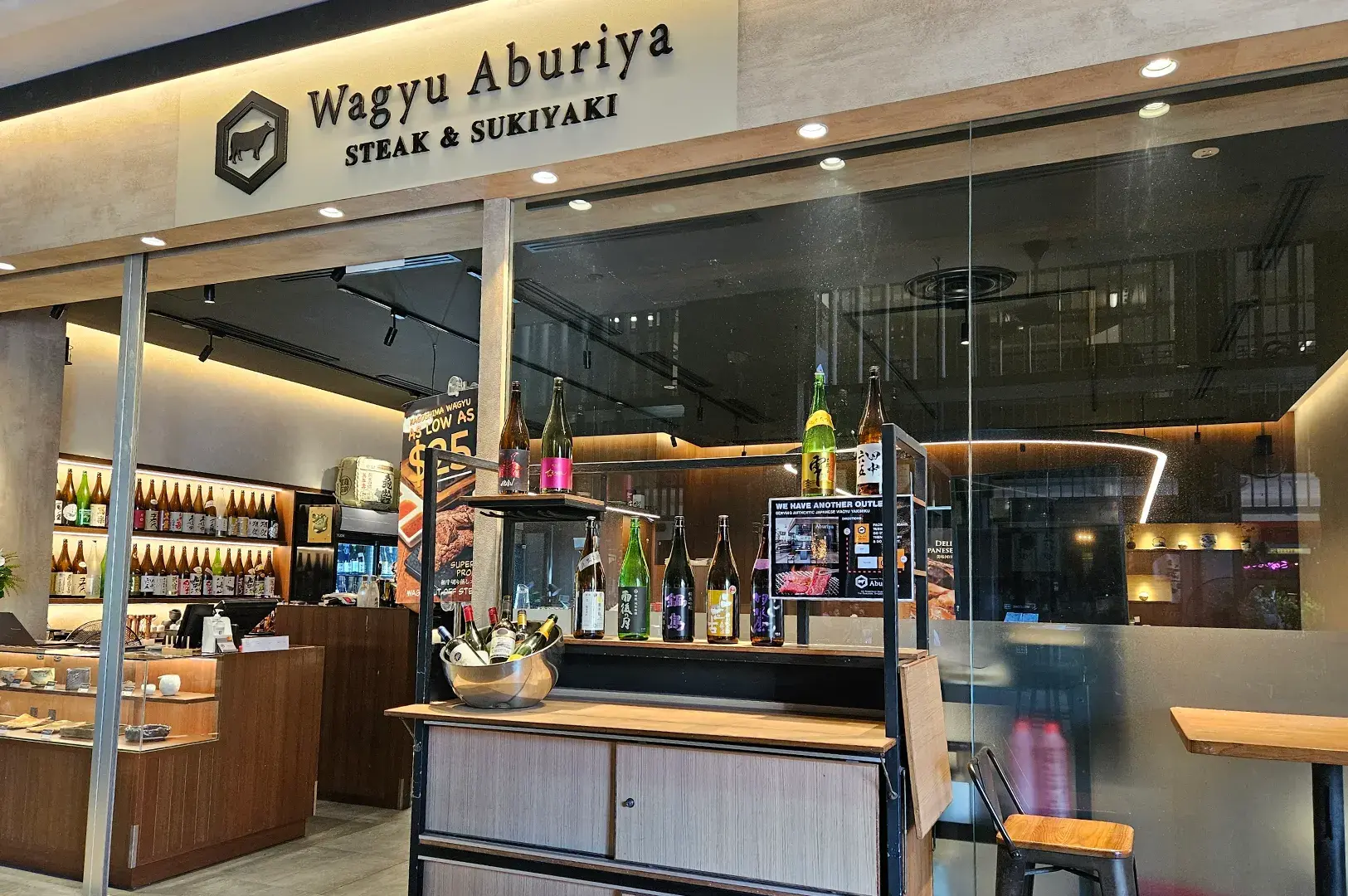 Interior entry view of Wagyu Aburiya Steak & Sukiyaki showcasing a display shelf with sake bottles and modern dining seating.