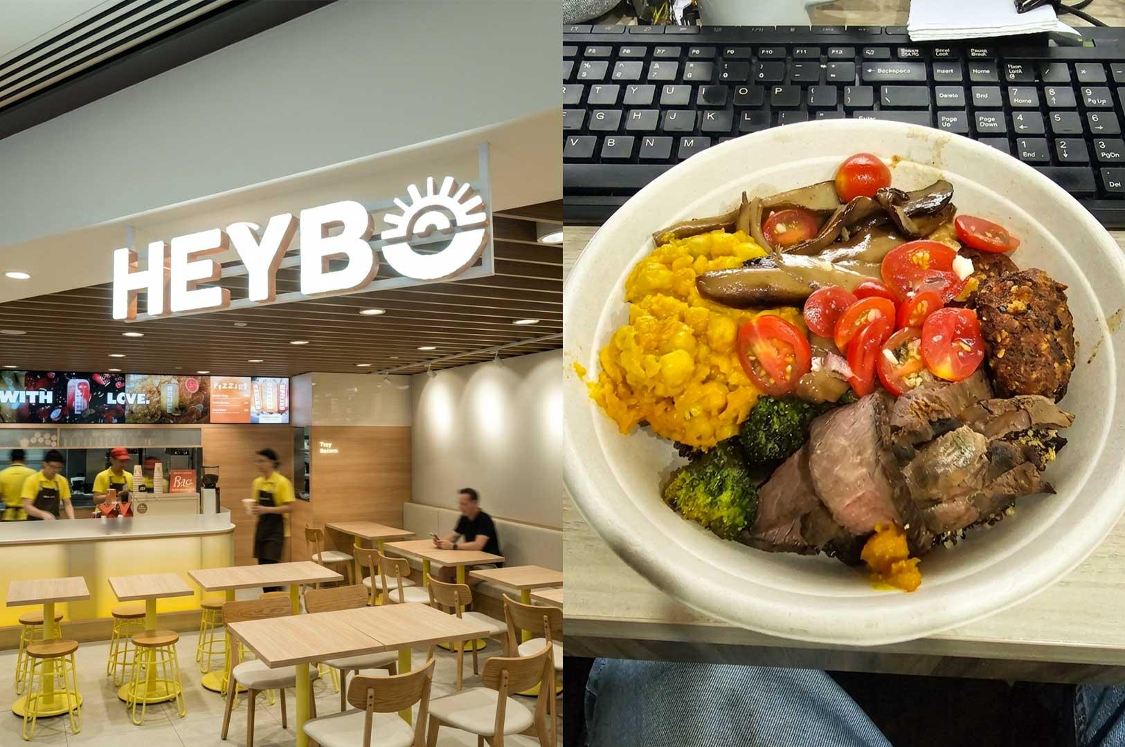 Left Panel: The bright, modern storefront and seating area of "HEYBO," featuring a large illuminated brand sign, warm wooden dining tables paired with yellow metal stools and light-wood chairs, modern overhead recessed lighting, and staff in yellow aprons behind the counter. Right Panel: A top-down shot of a takeaway grain bowl resting on an office desk next to a black computer keyboard, filled with sliced roasted steak, halved cherry tomatoes, sauteed mushrooms, charred broccoli florets, yellow turmeric grain/corn mix, and a savory veggie patty.
