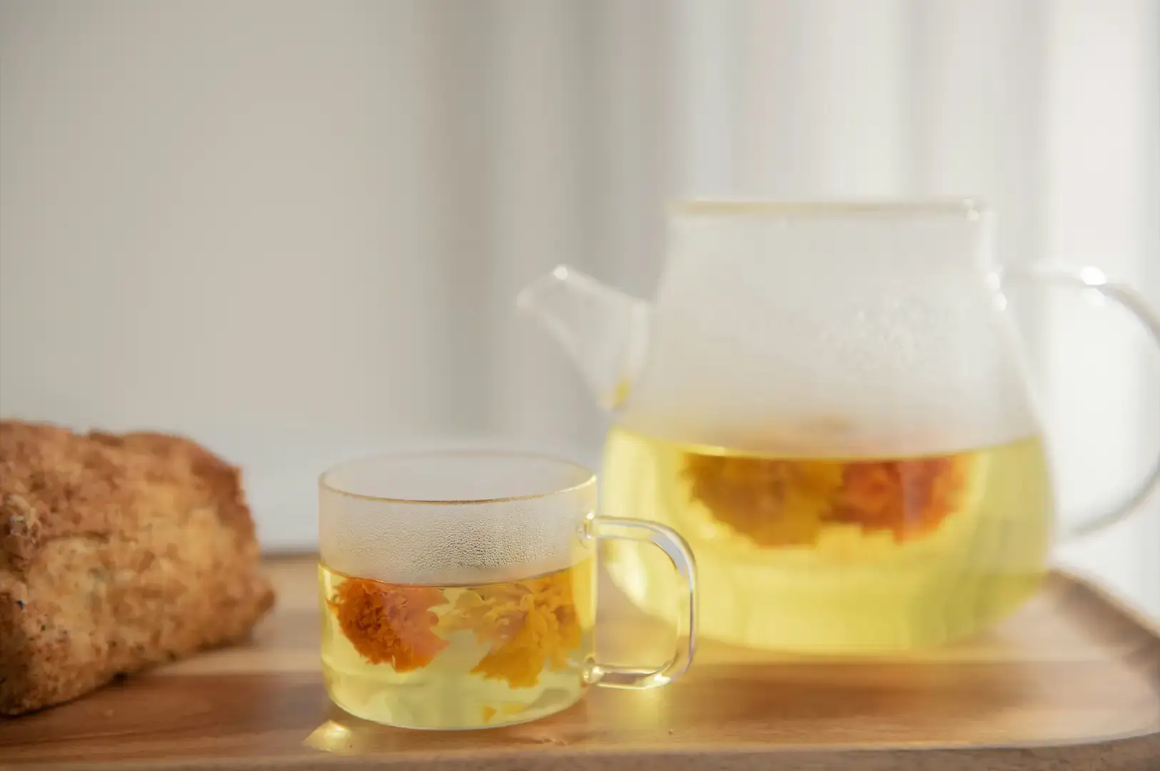 A bright and clean tabletop scene showcasing a clear glass teacup filled with pale yellow tea and blooming golden chrysanthemum flowers. Next to the cup stands a matching transparent glass teapot containing more of the steeping herbal infusion. The set is arranged on a light-colored wooden serving tray, with a crusty baked pastry or scone partially visible on the left side against a soft, bright, neutral background.