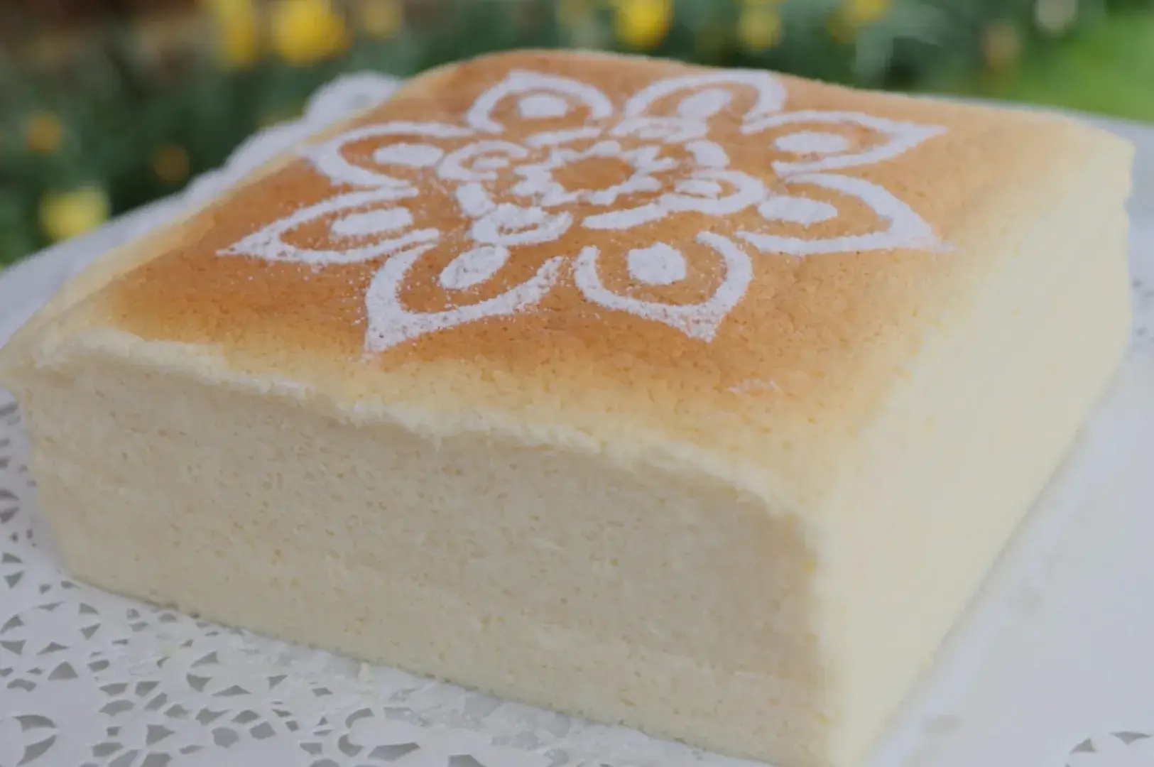 A square, fluffy cake topped with a decorative flower pattern made of powdered sugar on a lace doily.