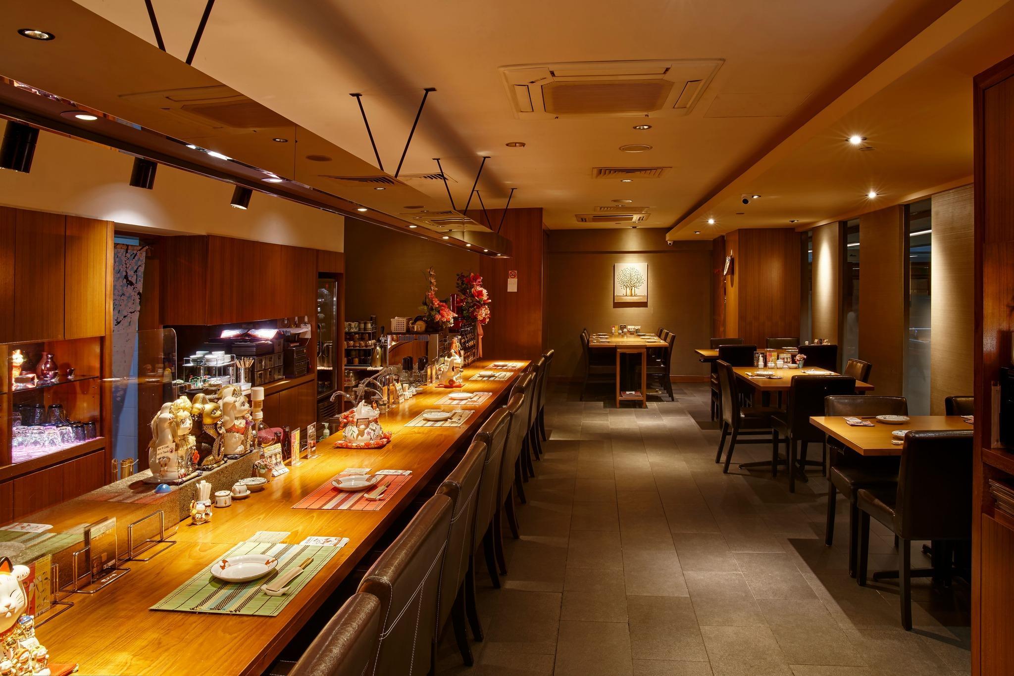 The image showcases a warm, inviting restaurant interior featuring a long, polished wooden counter lined with Maneki-neko figurines. The space is elegantly lit with soft overhead lamps and track lighting, highlighting several dining tables set for service in the background.