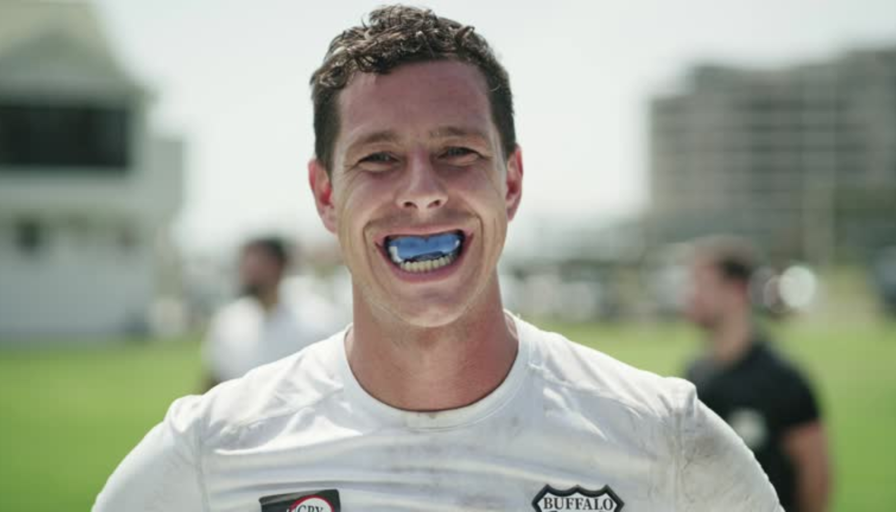 "Mann i hvit t-skjorte smiler med blått tannvern i munnen, utendørs på sportsbane."