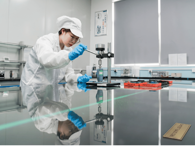 A laboratory technician in a white cleanroom suit, cap, and blue gloves using a precision measurement instrument on a highly reflective black table. The background shows a professional, well-lit laboratory environment.