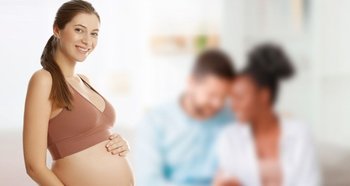 Surrogate holding her pregnant belly with the intended parents blurred behind her
