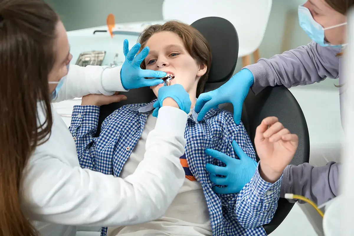 A dental procedure on a young patient in a blue checkered shirt, with dental professionals in blue gloves examining their mouth