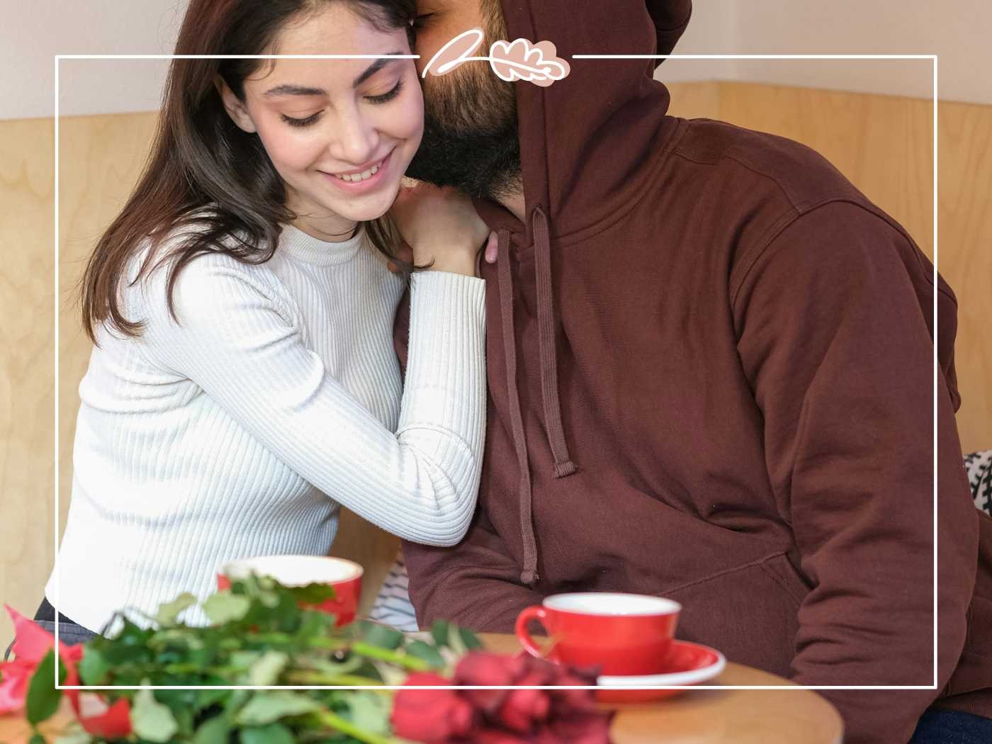 A cozy moment between a smiling woman and a bearded man, sharing an affectionate hug at a table with a red coffee cup and red roses - Fabulous Flowers and Gifts.