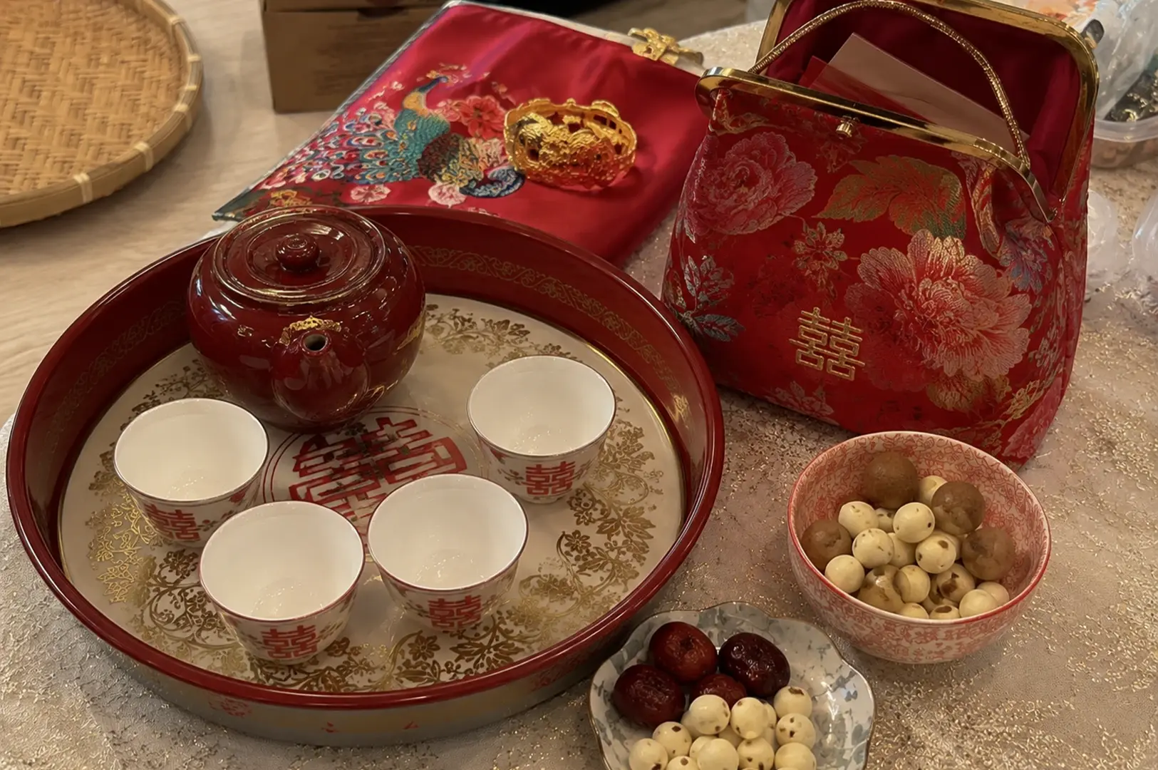 A collection of traditional Chinese wedding items is arranged on a glittering, light-gold tablecloth. In the foreground, a large round red lacquer tray features a glossy red teapot and four small white ceramic teacups, each printed with the red "Double Happiness" character. Next to the tray, there is a small floral plate containing red dates and white lotus seeds, alongside a larger pink patterned bowl holding roasted nuts. In the background, a matching red silk handbag with gold floral embroidery and the "Double Happiness" symbol sits near a folded red silk fabric decorated with a colorful embroidered peacock and a thick gold dragon bangle.