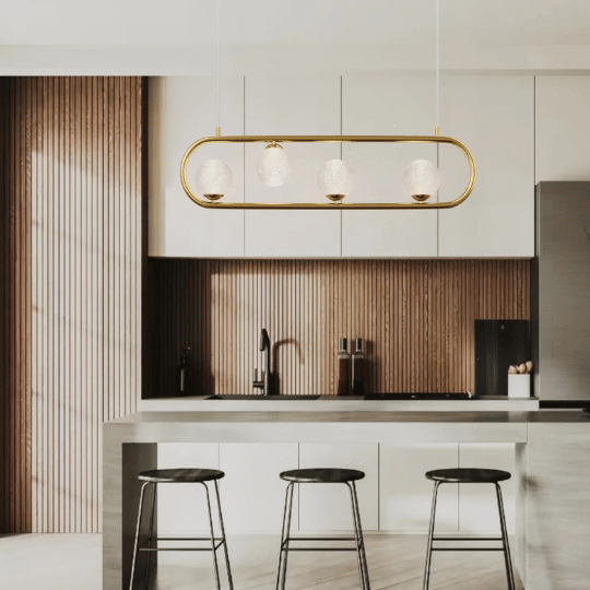 Modern kitchen with a concrete island and black barstools, illuminated by a gold linear pendant light featuring four spherical glass shades.