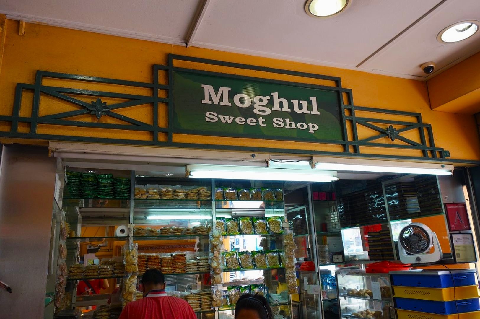 Exterior of Moghul Sweet Shop with a green sign. Shelves with colorful packets and snacks are visible through the glass. A warm, inviting atmosphere.