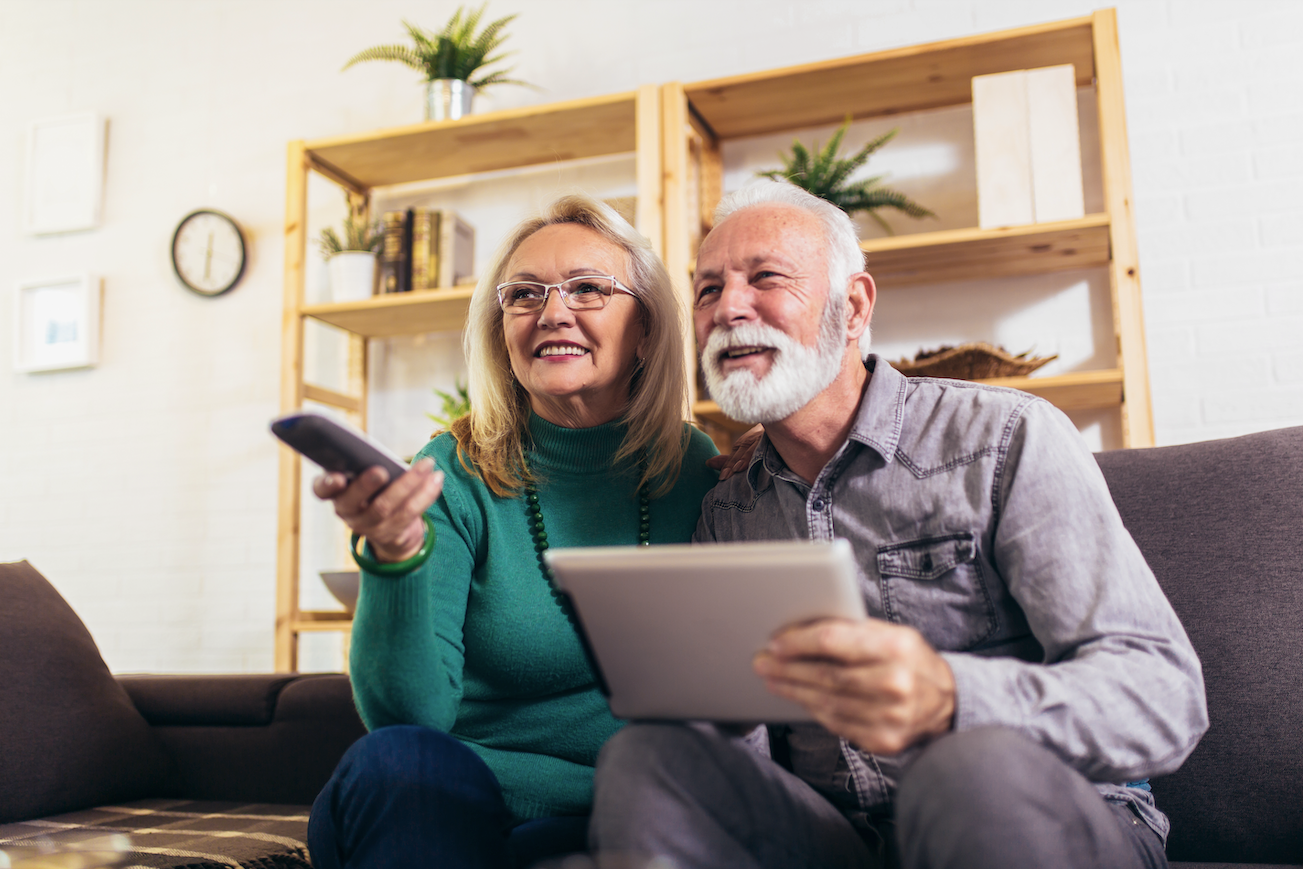 an older couple sit on the couch with a tablet and tv remote in their hands