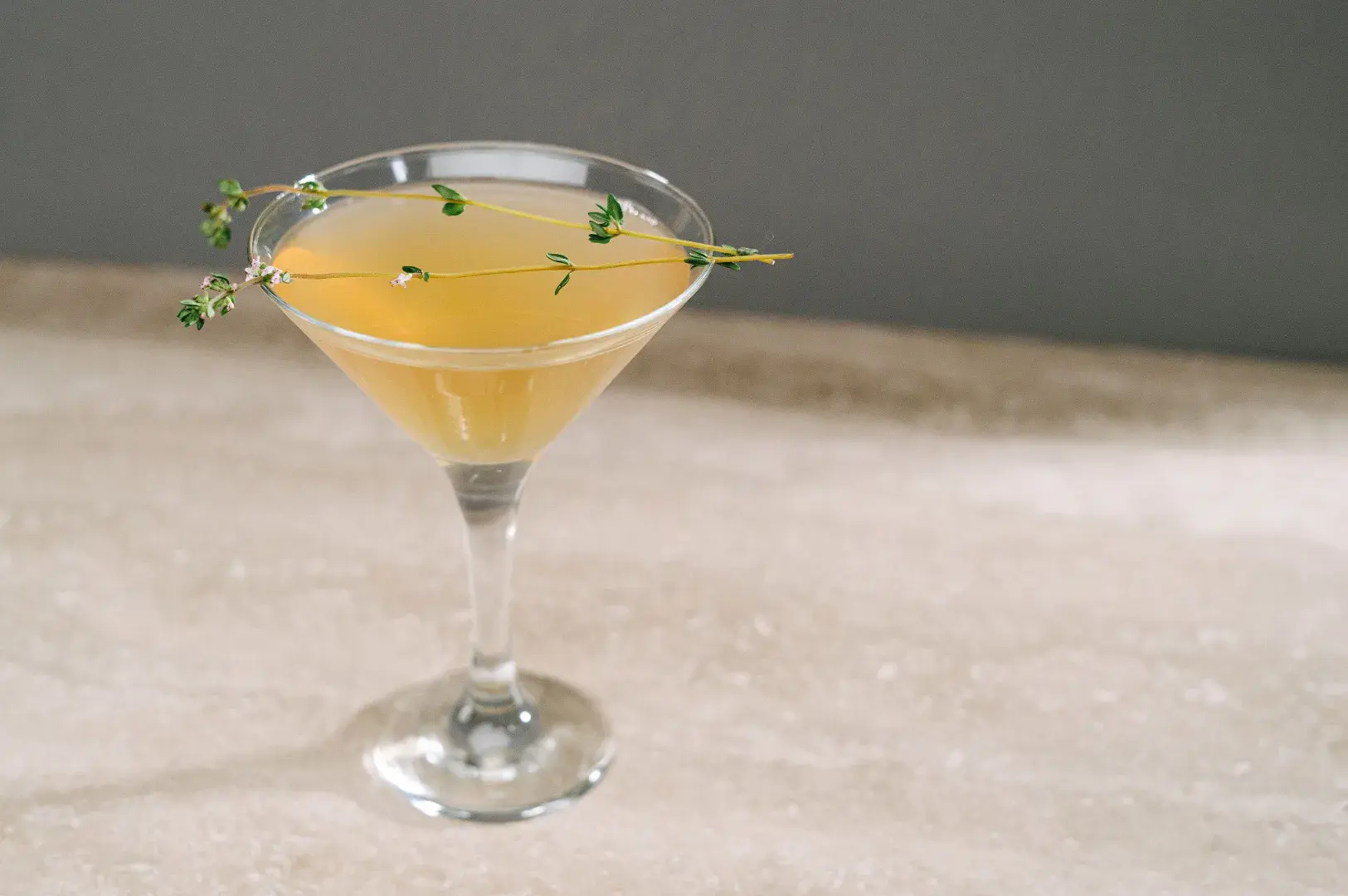 Elegant martini glass filled with a light amber cocktail and garnished with sprigs of fresh green thyme resting across the rim.