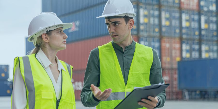 Two workers in safety vests and helmets discussing logistics at a shipping container yard.