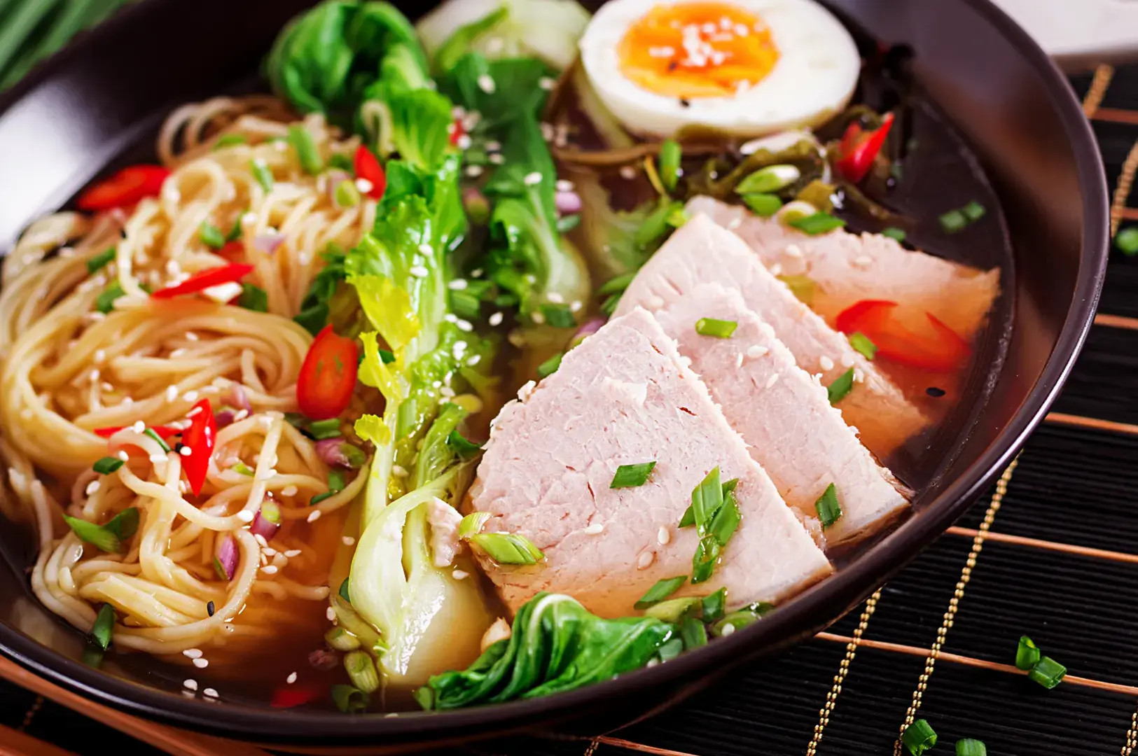 A bowl of ramen with noodles, sliced pork, bok choy, a hard-boiled egg, chili slices, and sesame seeds.