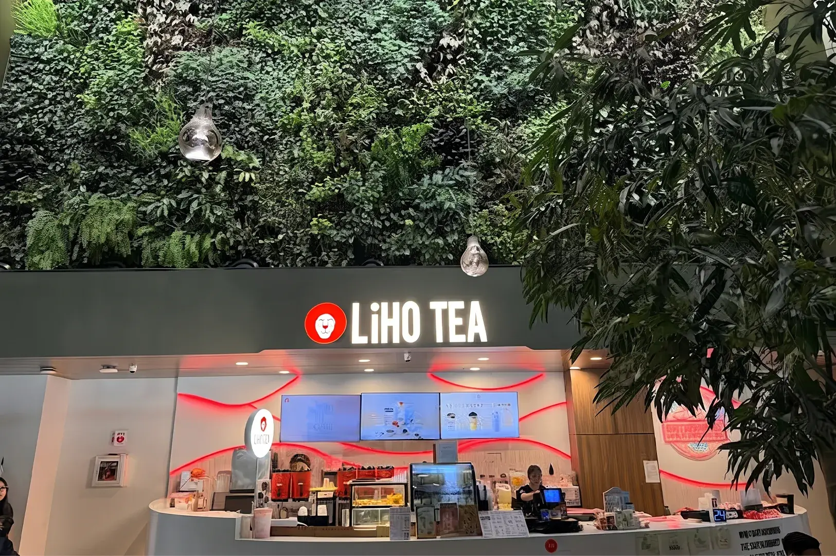 Storefront of a LiHO TEA outlet set against a lush green vertical garden wall, with white illuminated signage and red LED wave accents behind the ordering counter.