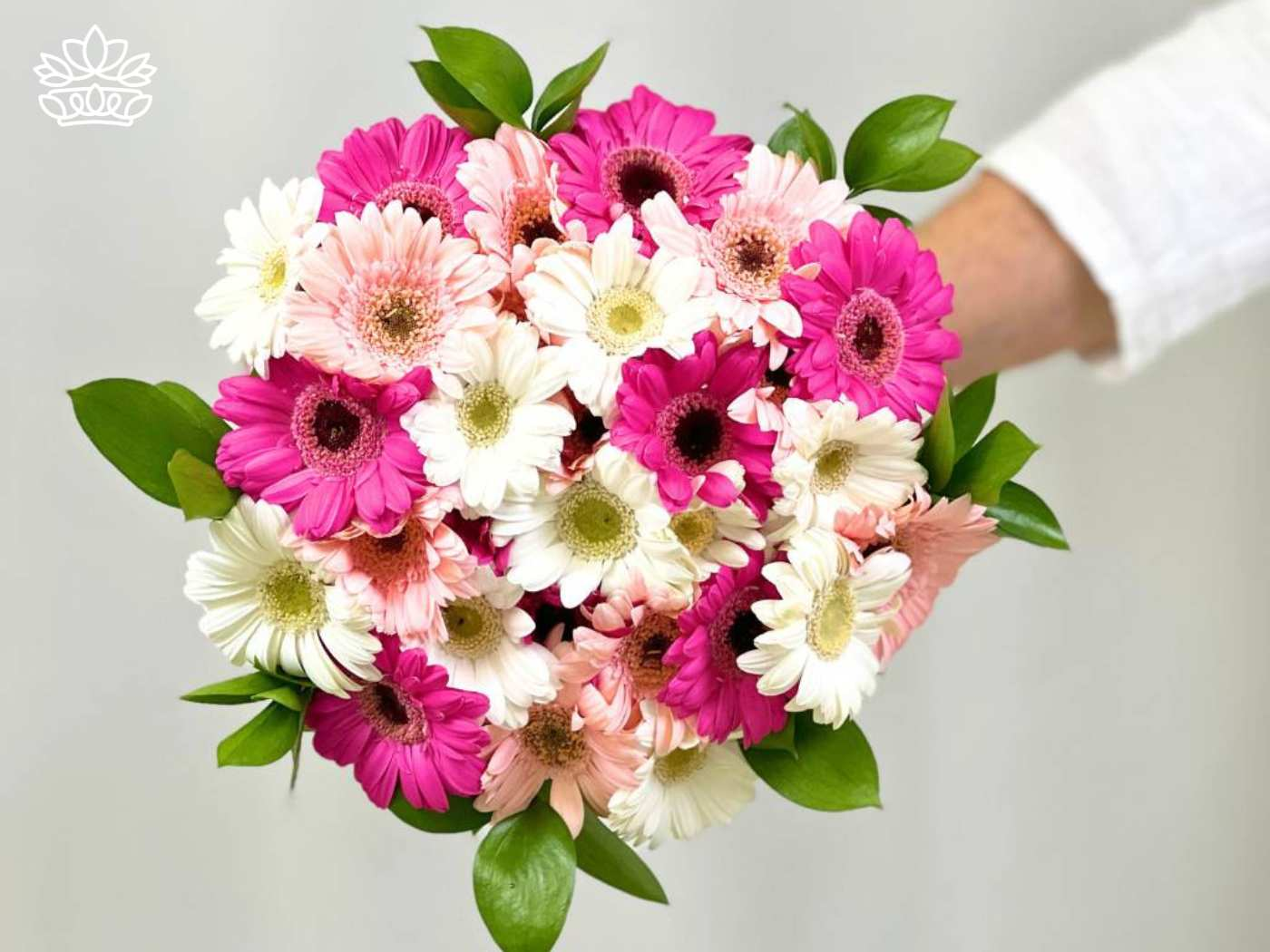 A bouquet of pink, white, and light peach Gerbera daisies, complemented by green foliage and held by a person in a white sleeve. Fabulous Flowers and Gifts. Gerberas Collection. Delivered with Heart.