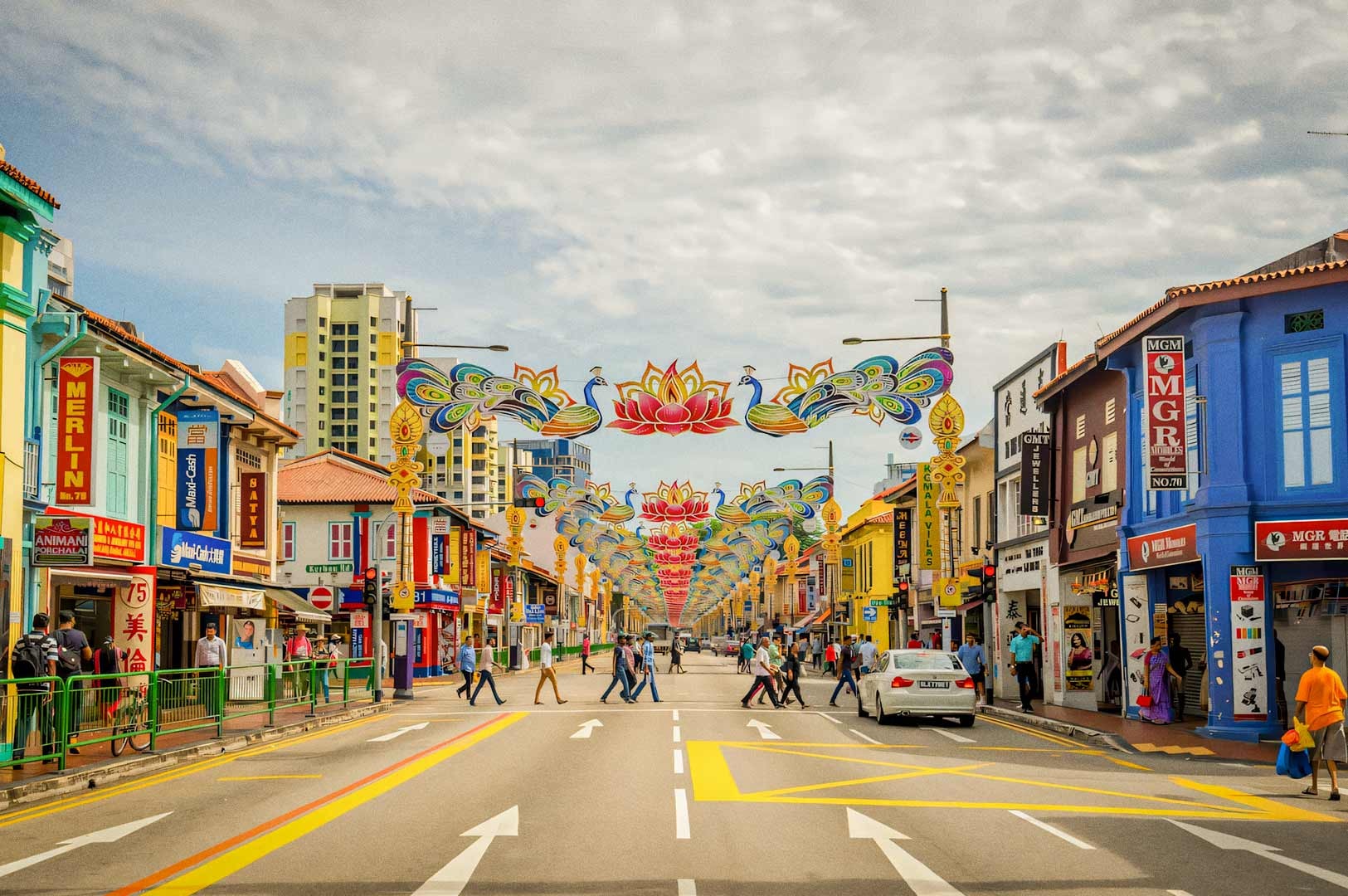 A colorful street scene lined with vibrant shophouses and decorated with elaborate floral and peacock-themed arches hanging over the road.