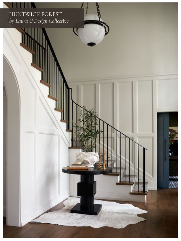 A classic white-paneled entryway with a sweeping staircase and black iron railings. A round black pedestal table with layered decor sits on a light hide rug, creating a strong central focal point.