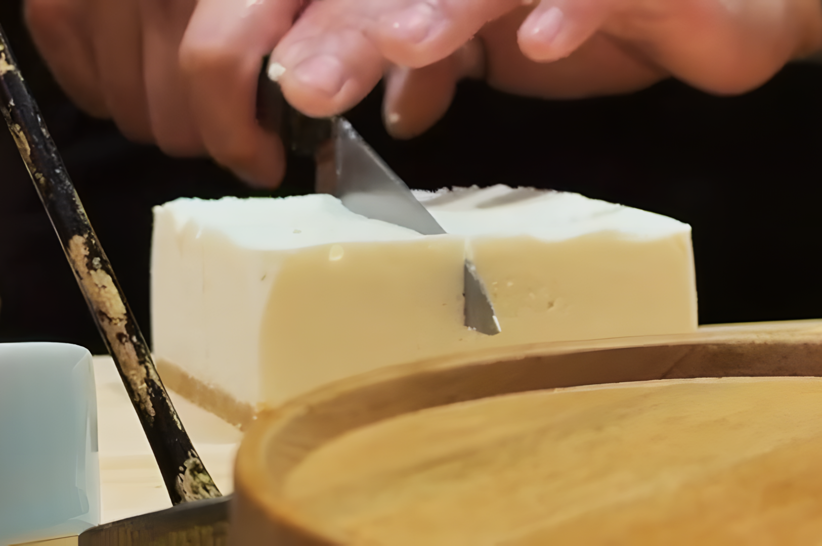 Hands carefully cutting a block of tofu on a wooden board. A knife slices smoothly through the soft texture, conveying precision and focus.