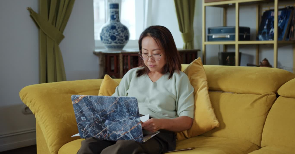 A woman sitting on a yellow couch, reviewing her schedule SE tax form on a laptop with focused attention.