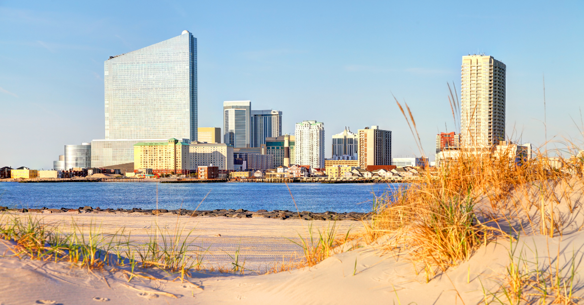 A scenic view of the Atlantic City skyline seen from the sandy beach with dune grass in the foreground and calm ocean water.
