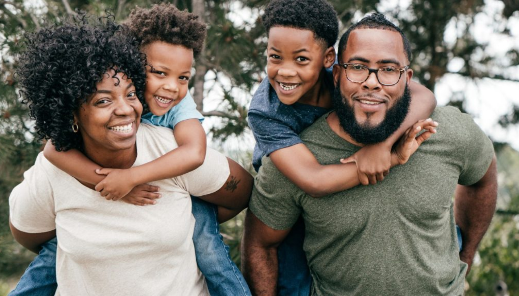 Smilende familie utendørs, to voksne bærer hvert sitt barn på ryggen mens de ser glade inn i kameraet.