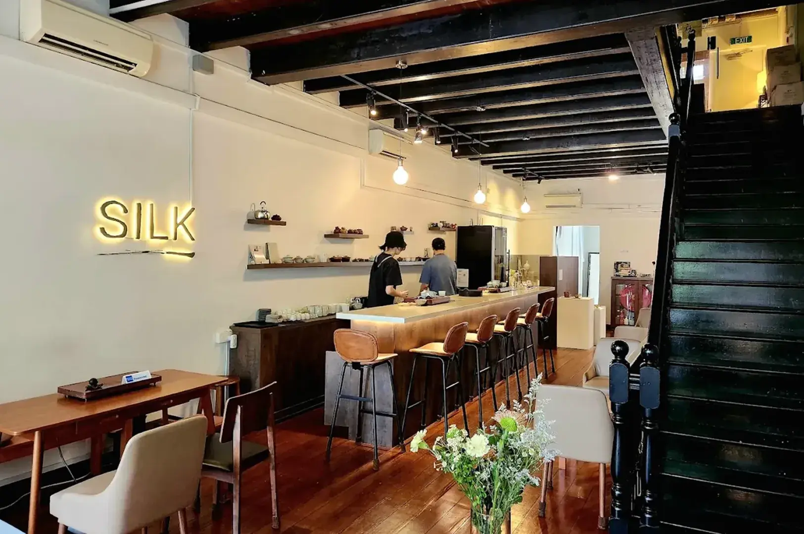 Interior of a modern teahouse featuring a illuminated "SILK" neon wall sign, counter seating with barstools, warm wooden floors, and dark ceiling beams.