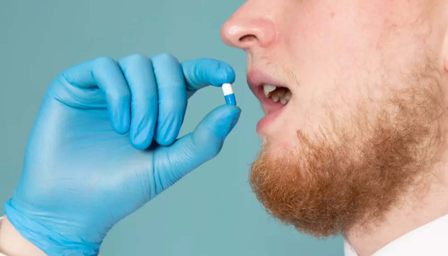 Mann med rød skjegg tar blå og hvit kapsel i munnen, holdt av hånd med blå hanske.